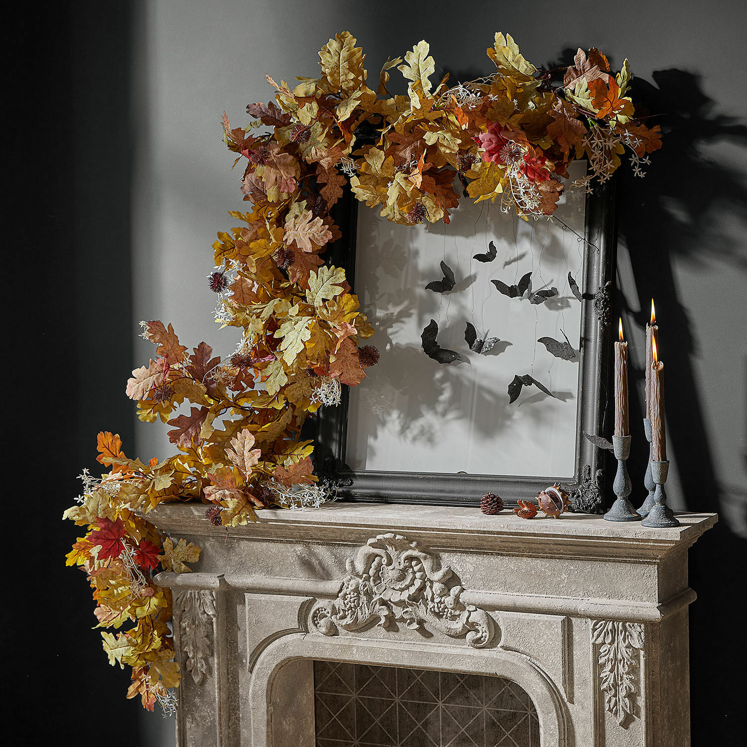 Guirnalda Fleurimont Corona decorativa otoñal de hojas de roble en tonos cálidos sobre chimenea de piedra con marco negro y velas