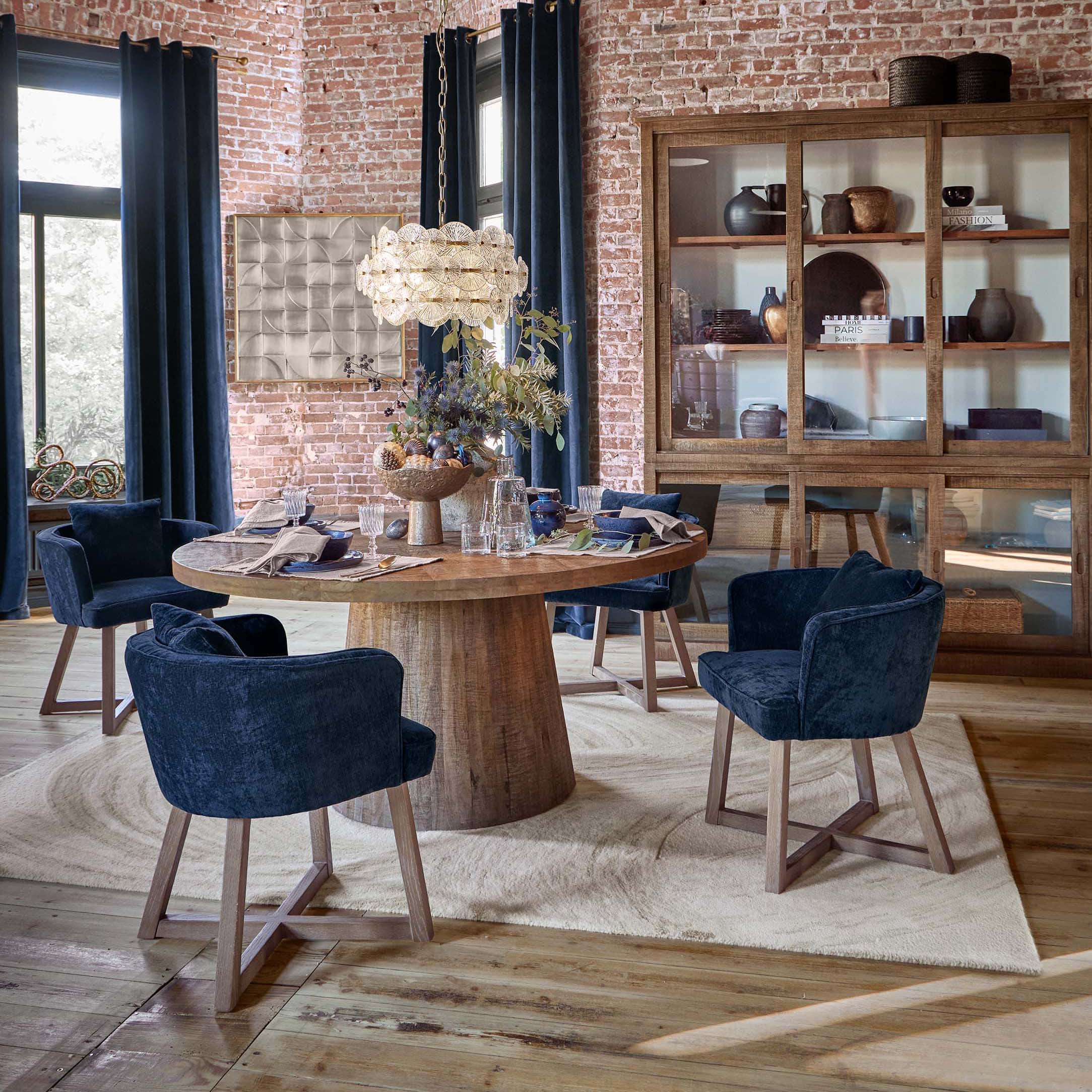 Comedor con mesa redonda de madera y sillas de terciopelo azul marino frente a pared de ladrillo y vitrina de madera