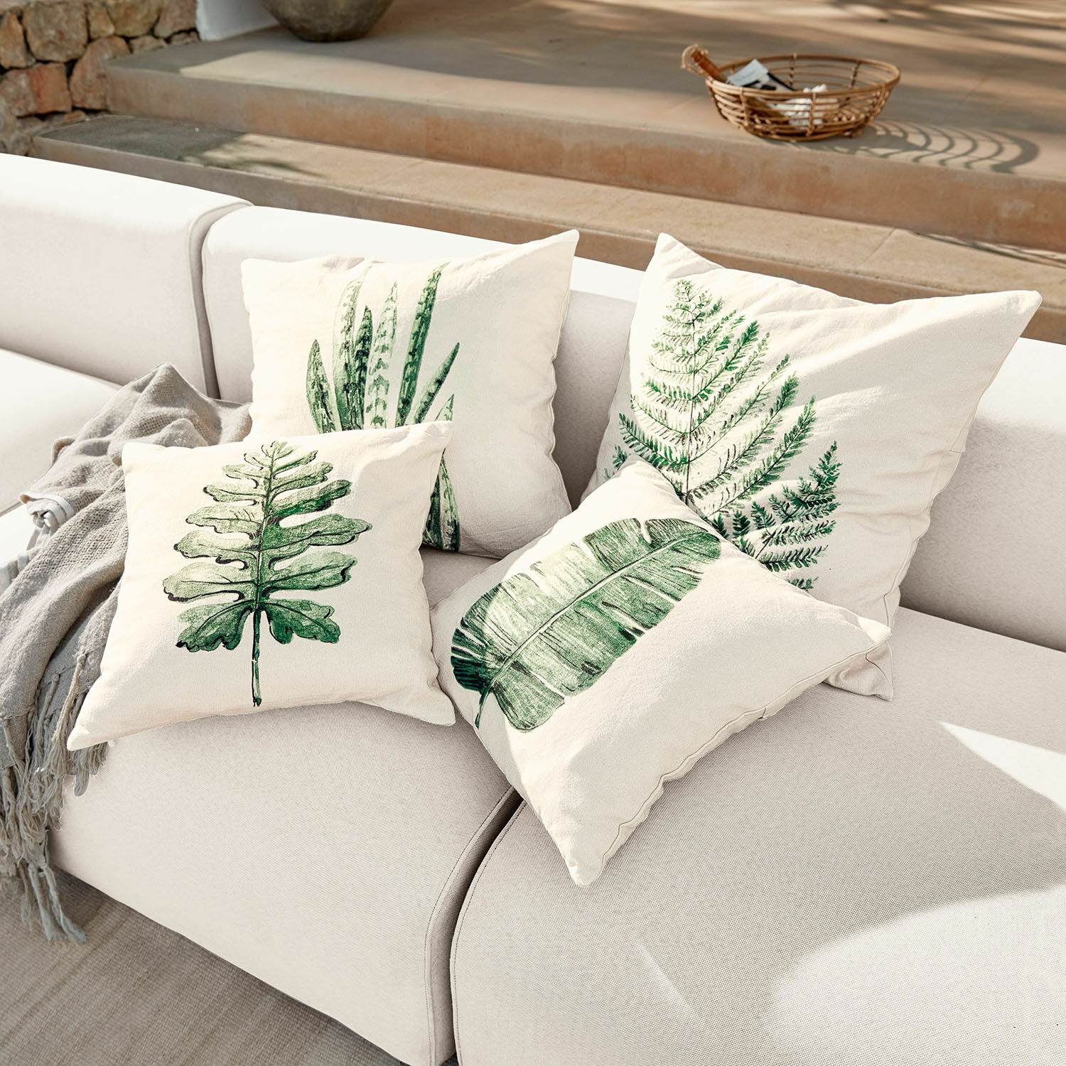Set de 4 fundas de cojín Marly Conjunto de cuatro cojines crema con estampados de hojas verdes sobre sofá claro, decoración interior