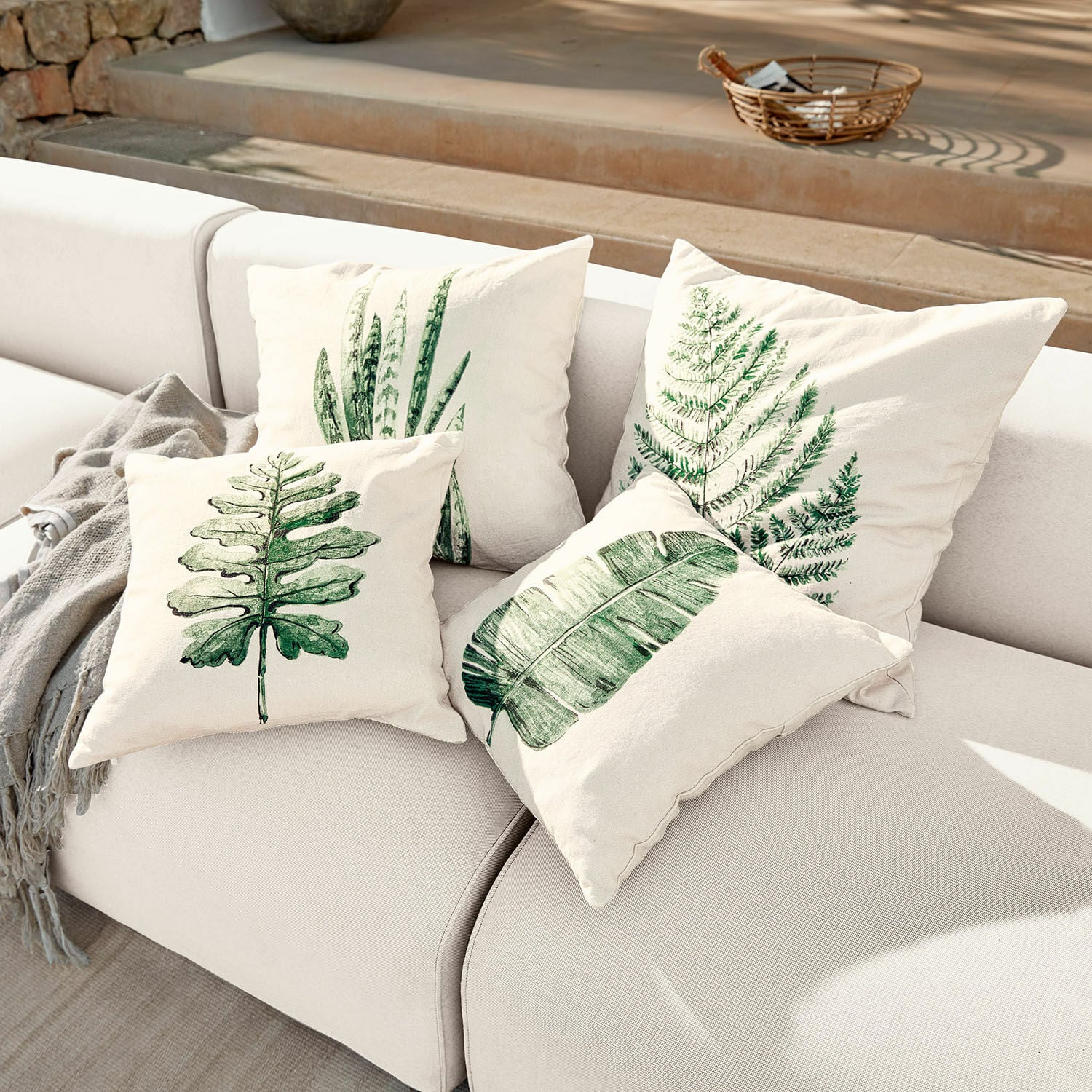 Set de 4 fundas de cojín Marly Conjunto de cuatro cojines crema con estampados de hojas verdes sobre sofá claro, decoración interior