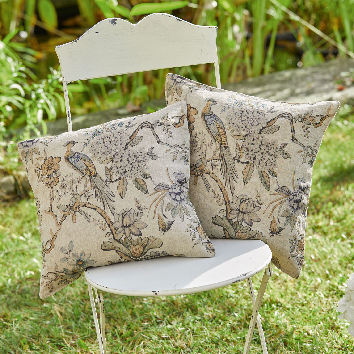 Dos cojines decorativos de algodón beige con estampado de pájaros y flores sobre una silla blanca en el jardín