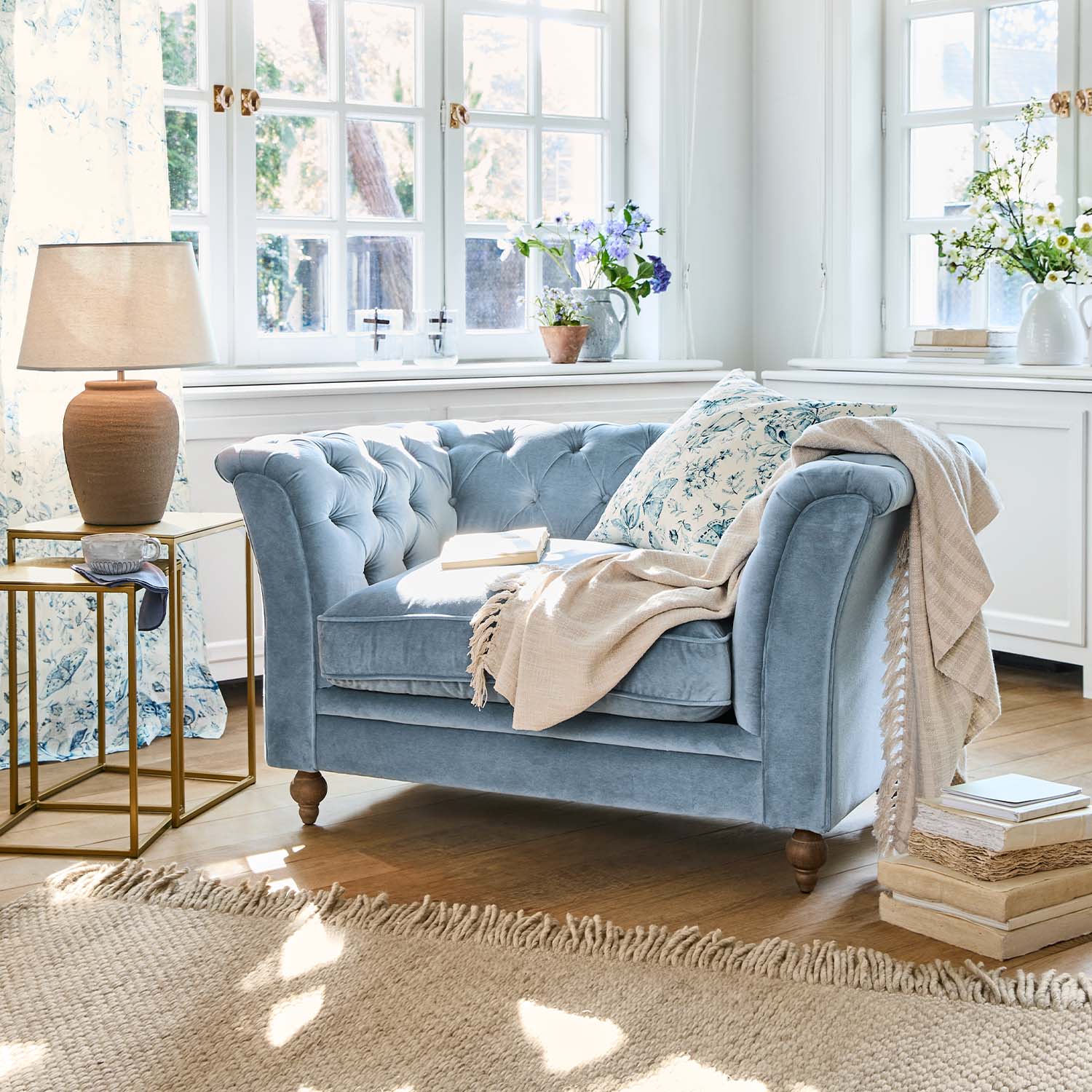 Sofá azul de terciopelo para dos personas con cojín floral y manta beige en salón luminoso con suelo de madera