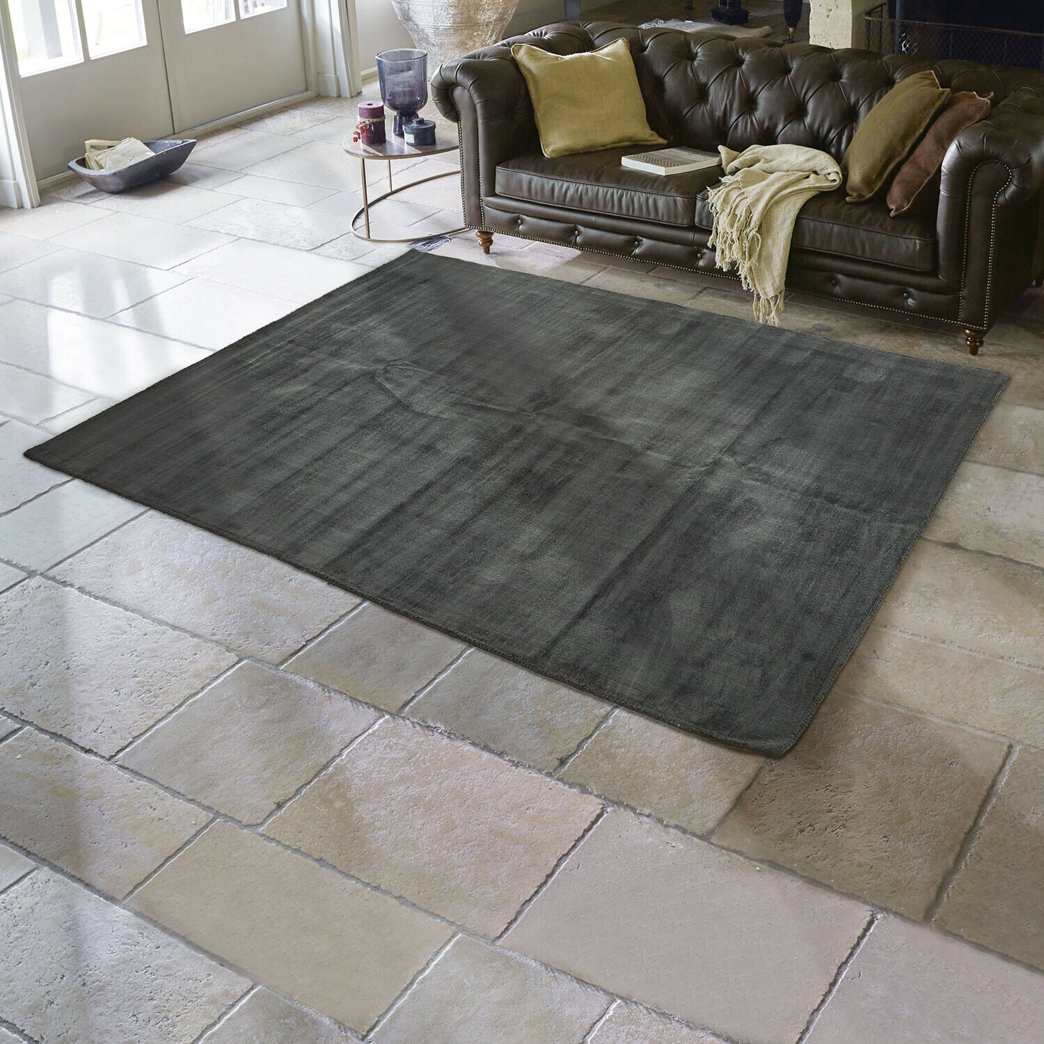 Alfombra Mariselle Alfombra gris oscuro de material suave sobre suelo de piedra clara en salón con sofá de cuero marrón