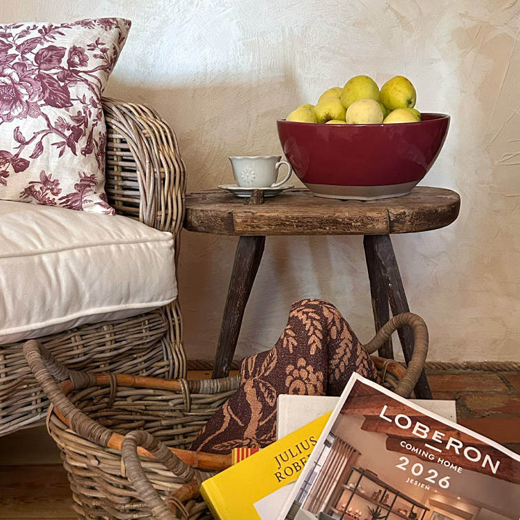 Mesa rústica de madera con cuenco cerámico burdeos lleno de manzanas amarillas junto a sillón de mimbre con cojín