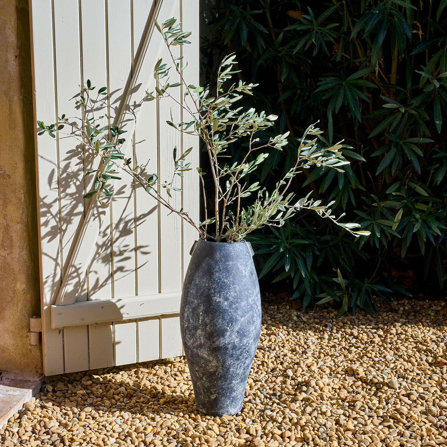 Jarrón alto gris de piedra con ramas de olivo sobre grava junto a puerta blanca