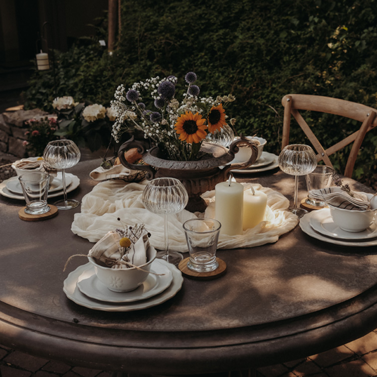 Mesa redonda de jardín con decoración de velas y flores en jarrón metálico para una reunión.
