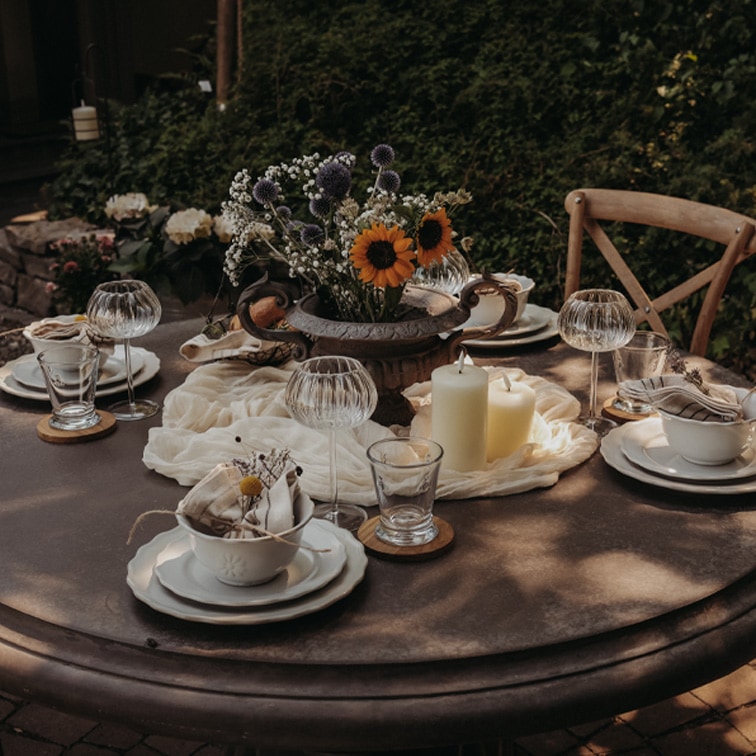 Mesa redonda de jardín con decoración de velas y flores en jarrón metálico para una reunión.