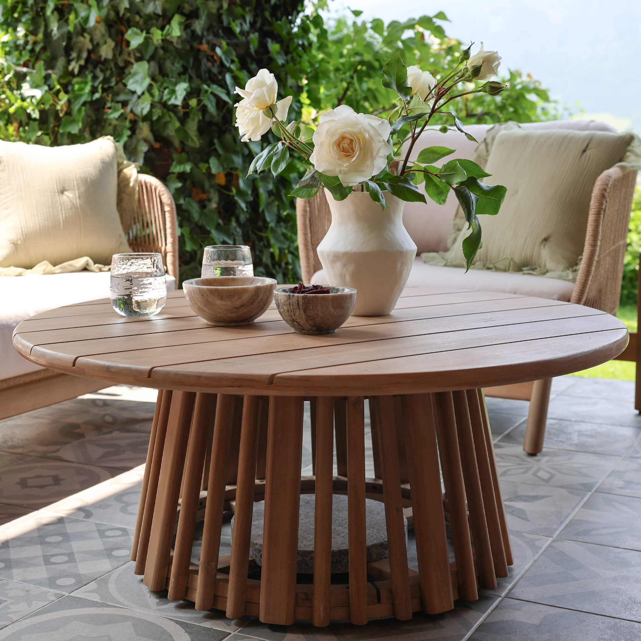 Mesa de centro redonda de madera de teca en color natural en terraza con decoración floral y cuencos