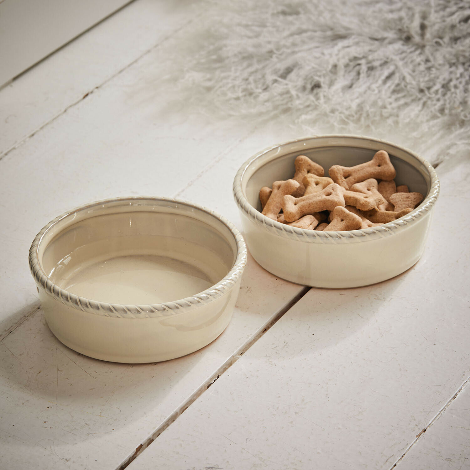 Set de 2 cuencos Pawilo Dos cuencos de cerámica crema para perros con borde decorativo sobre suelo de madera blanca