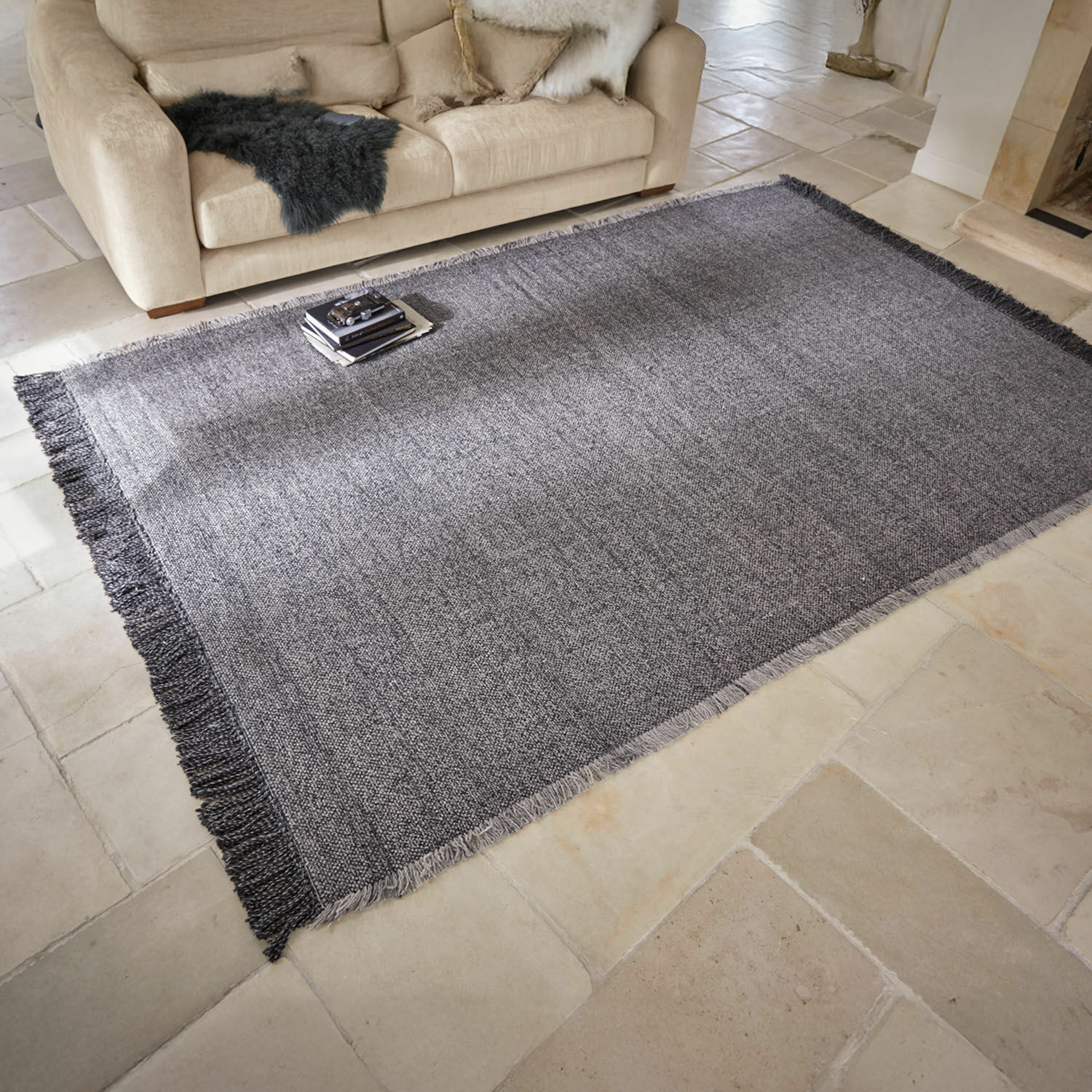 Alfombra para interior y exterior Marivela Alfombra gris con flecos sobre suelo claro en salón con sofá beige y cojines decorativos