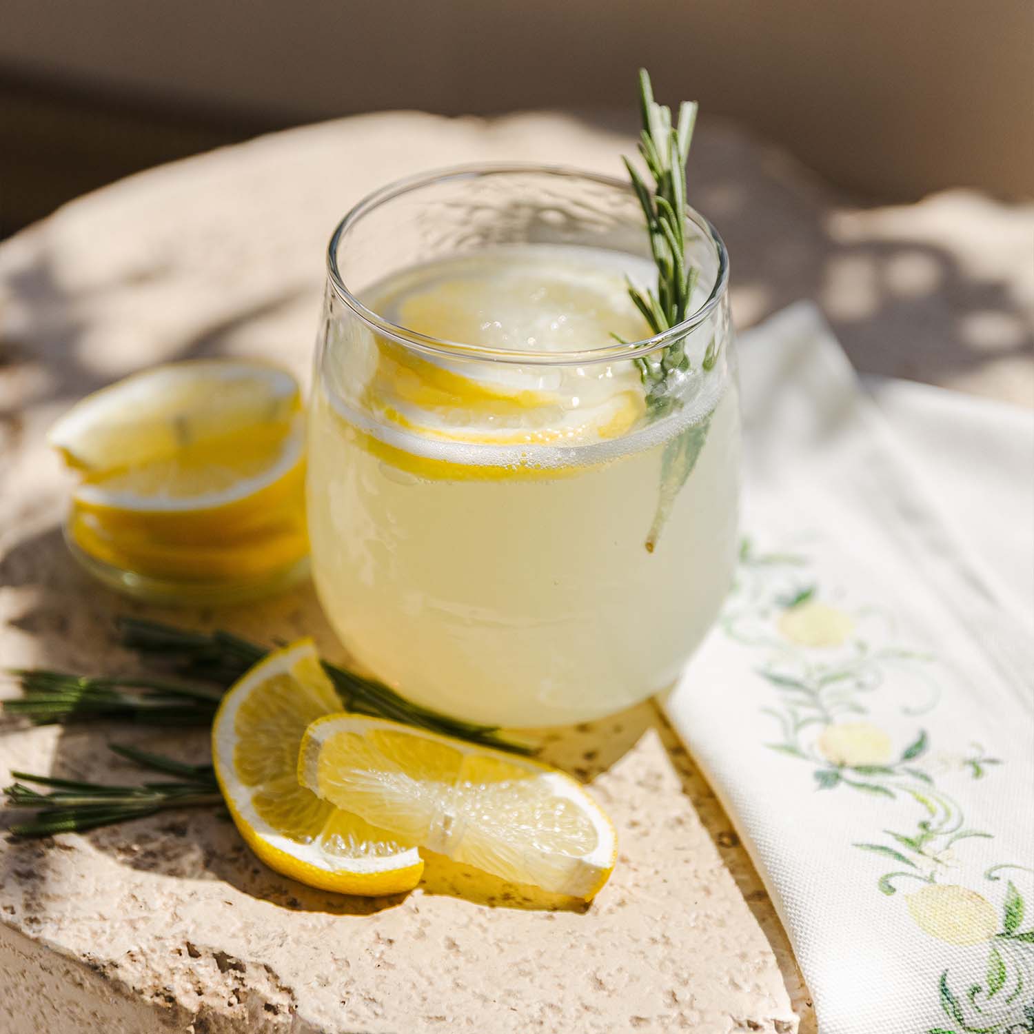 Vaso de limonada con rodajas de limón y ramita de romero sobre superficie de piedra con servilleta