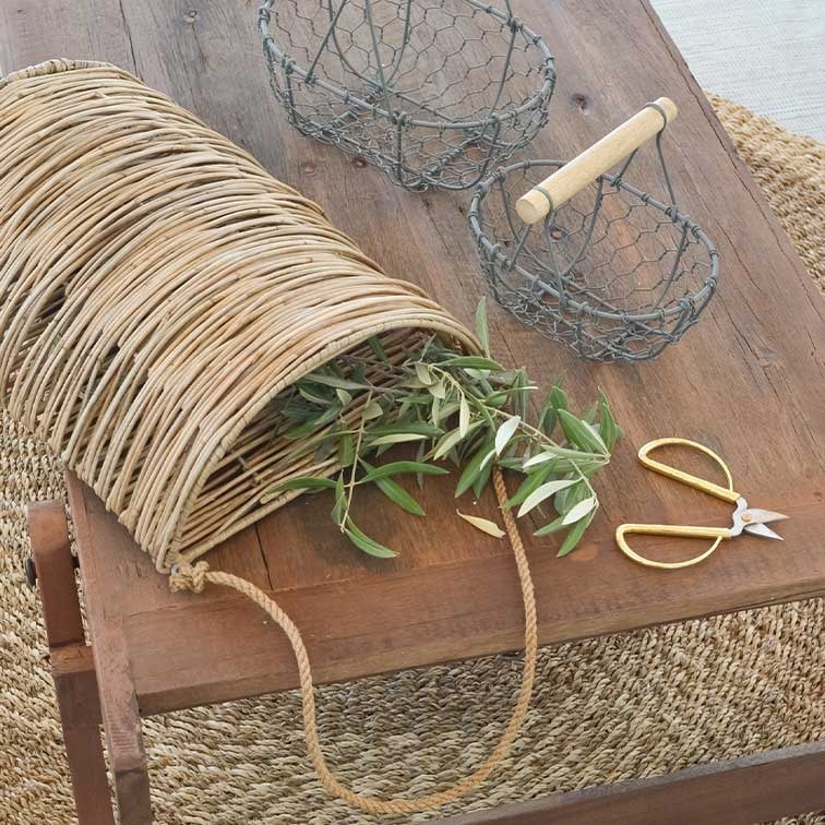 Cesta de mimbre con asa de cuerda sobre mesa de madera con ramas verdes y tijeras de jardín doradas