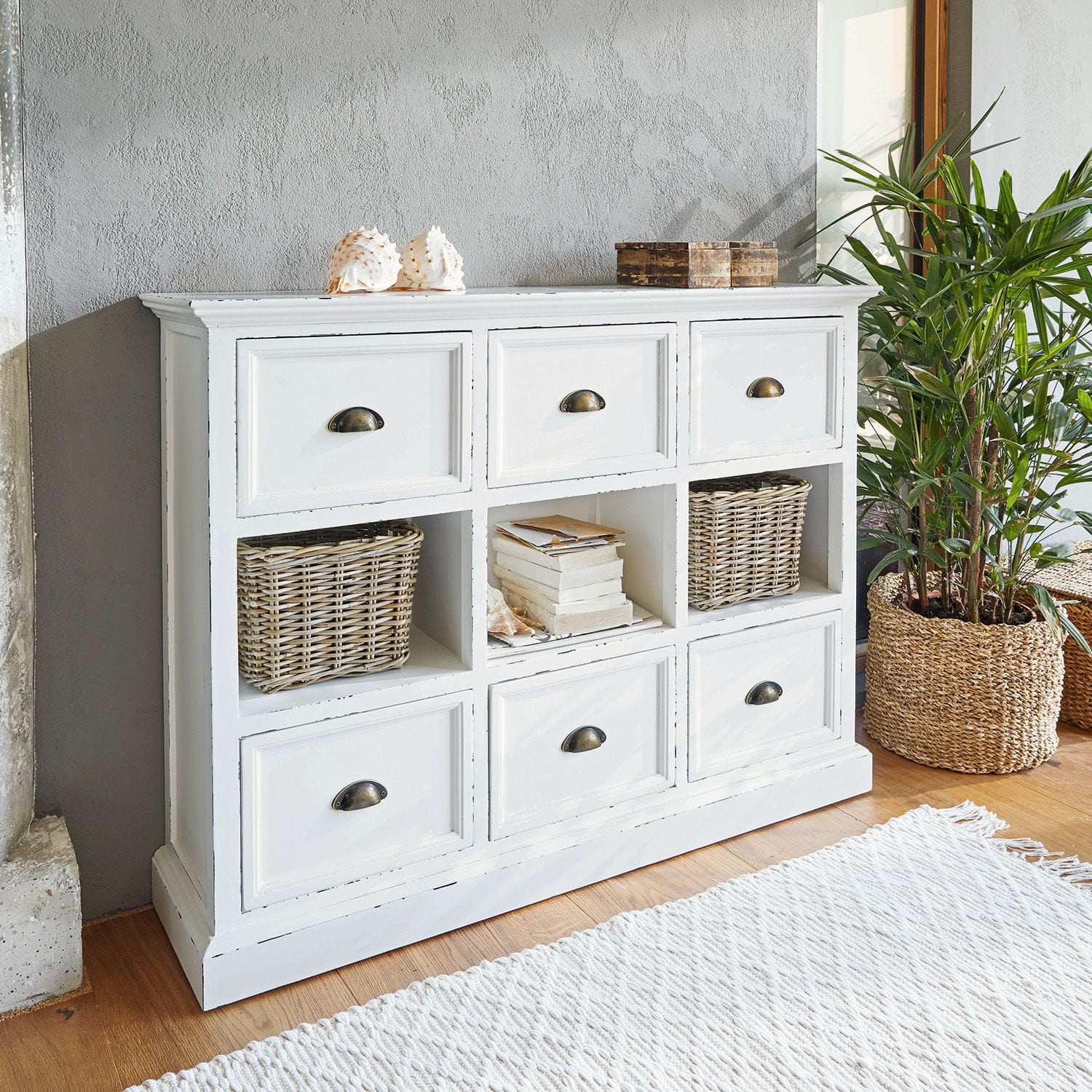 Cómoda Remilly Cómoda blanca de madera con seis cajones y tres estantes abiertos, decoraciones y planta al lado
