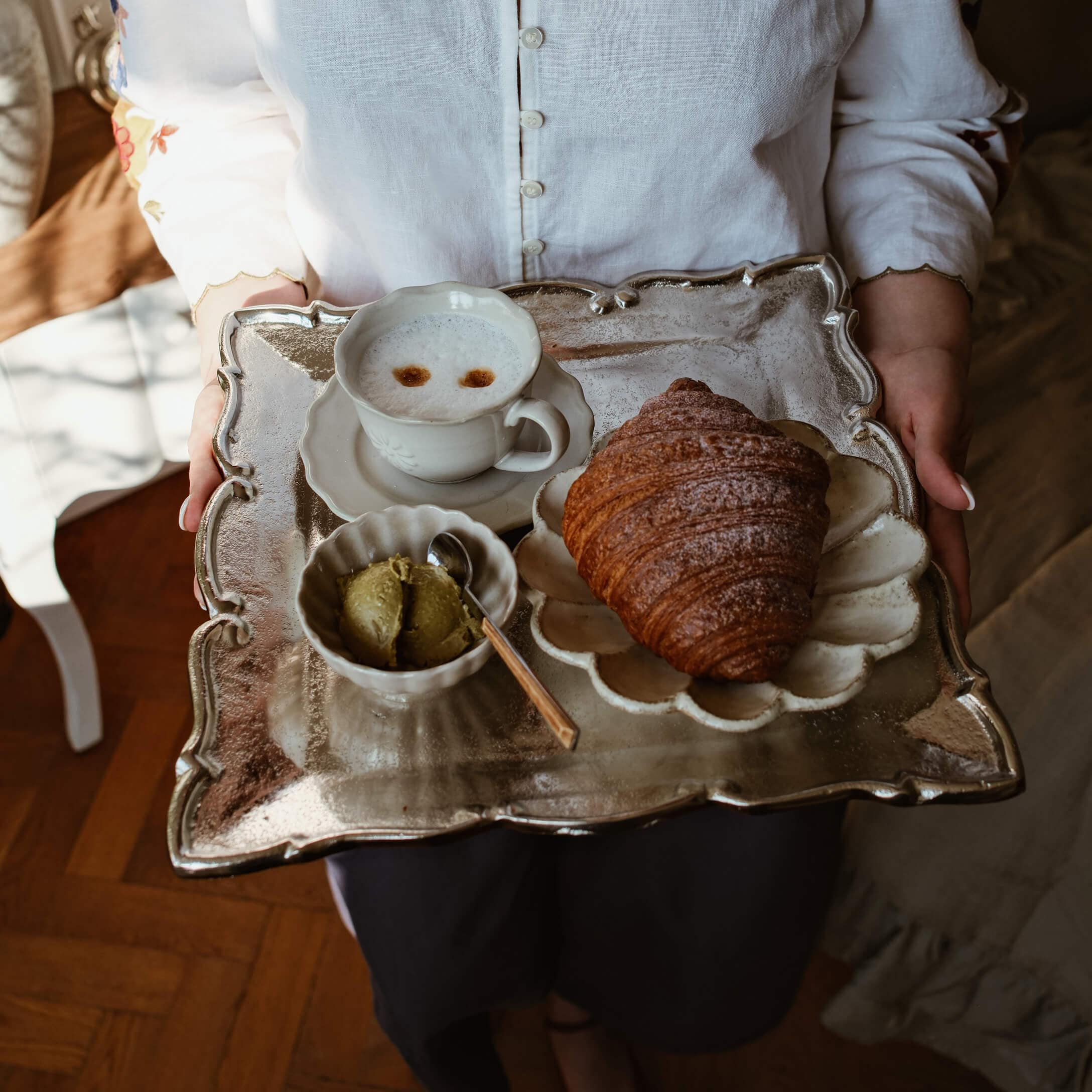 Bandeja metálica con croissant, taza de café y mantequilla verde sobre porcelana blanca