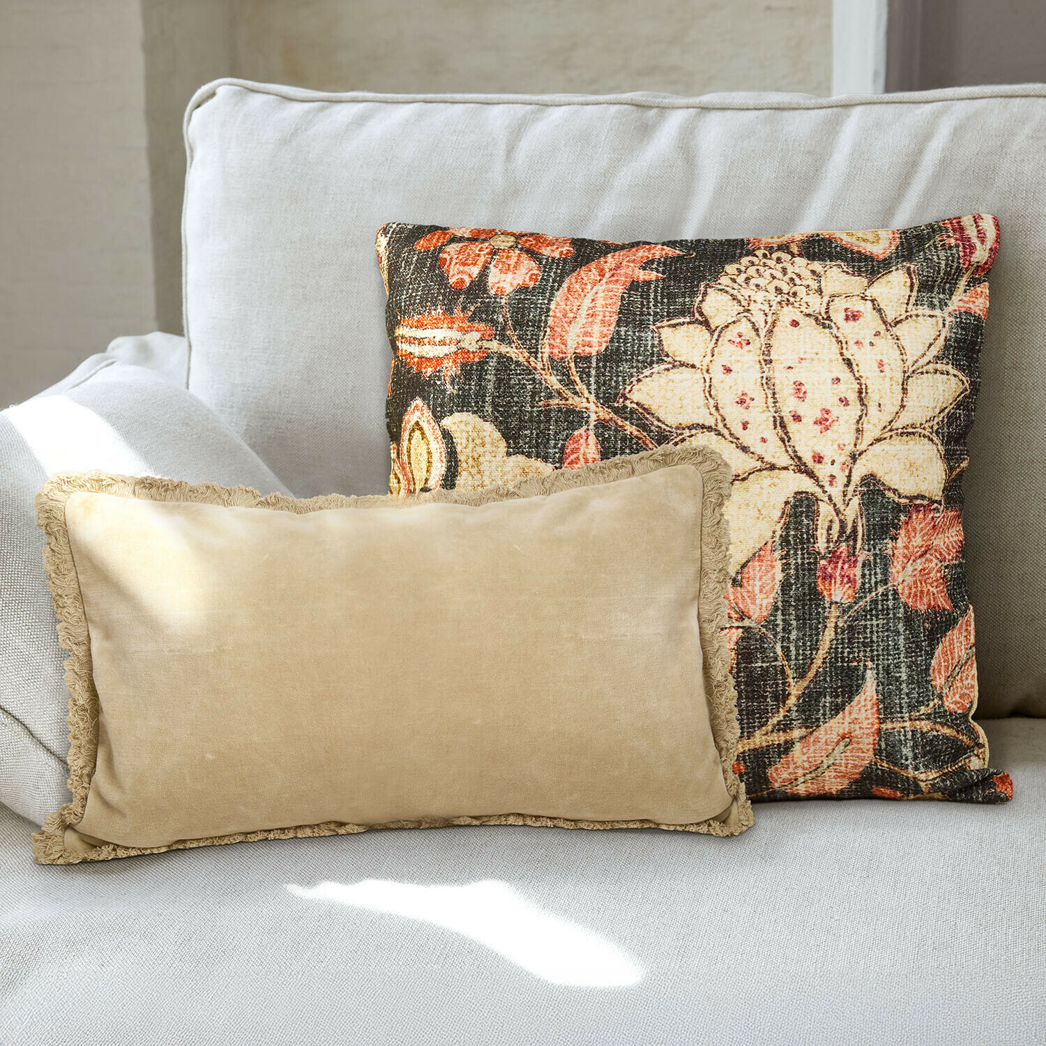 Set de 2 fundas de cojín Ambrayelle Dos cojines decorativos sobre un sofá gris claro, uno beige con flecos y otro con diseño floral colorido