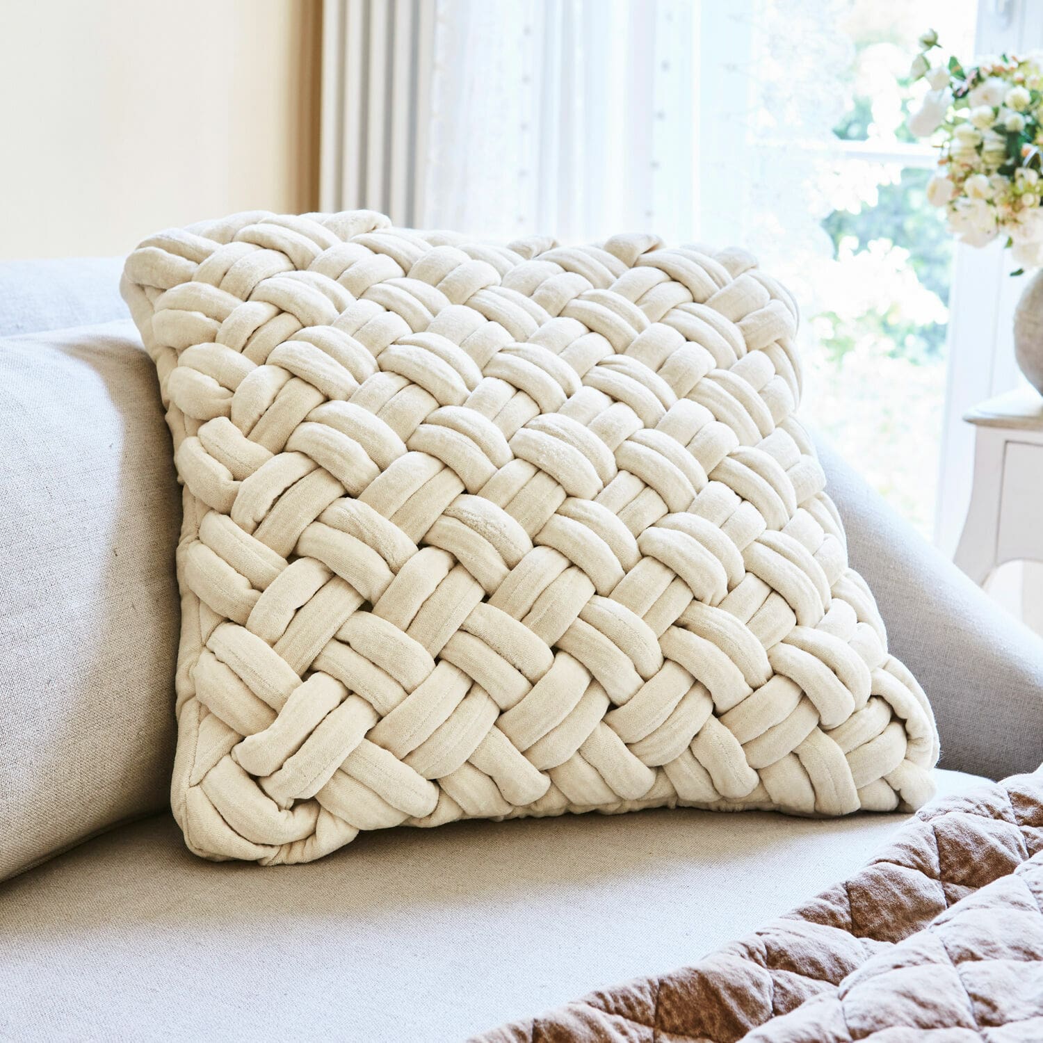 Cojín decorativo beige de material suave con diseño trenzado sobre sofá en sala de estar