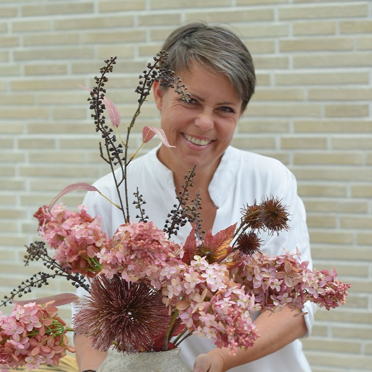 Mujer con blusa blanca sosteniendo un jarrón con flores secas decorativas rosas y marrones