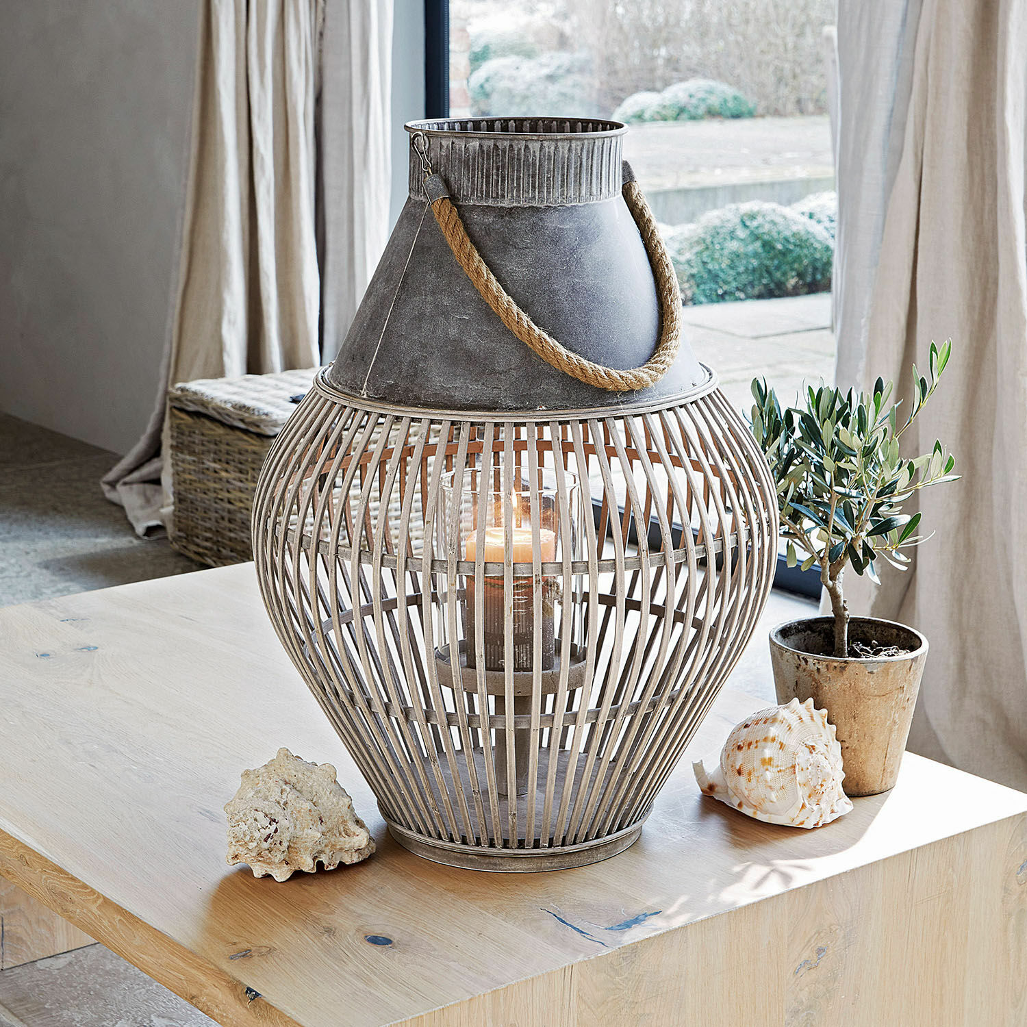 Linterna decorativa de metal y madera gris con asa de cuerda sobre mesa con conchas y planta