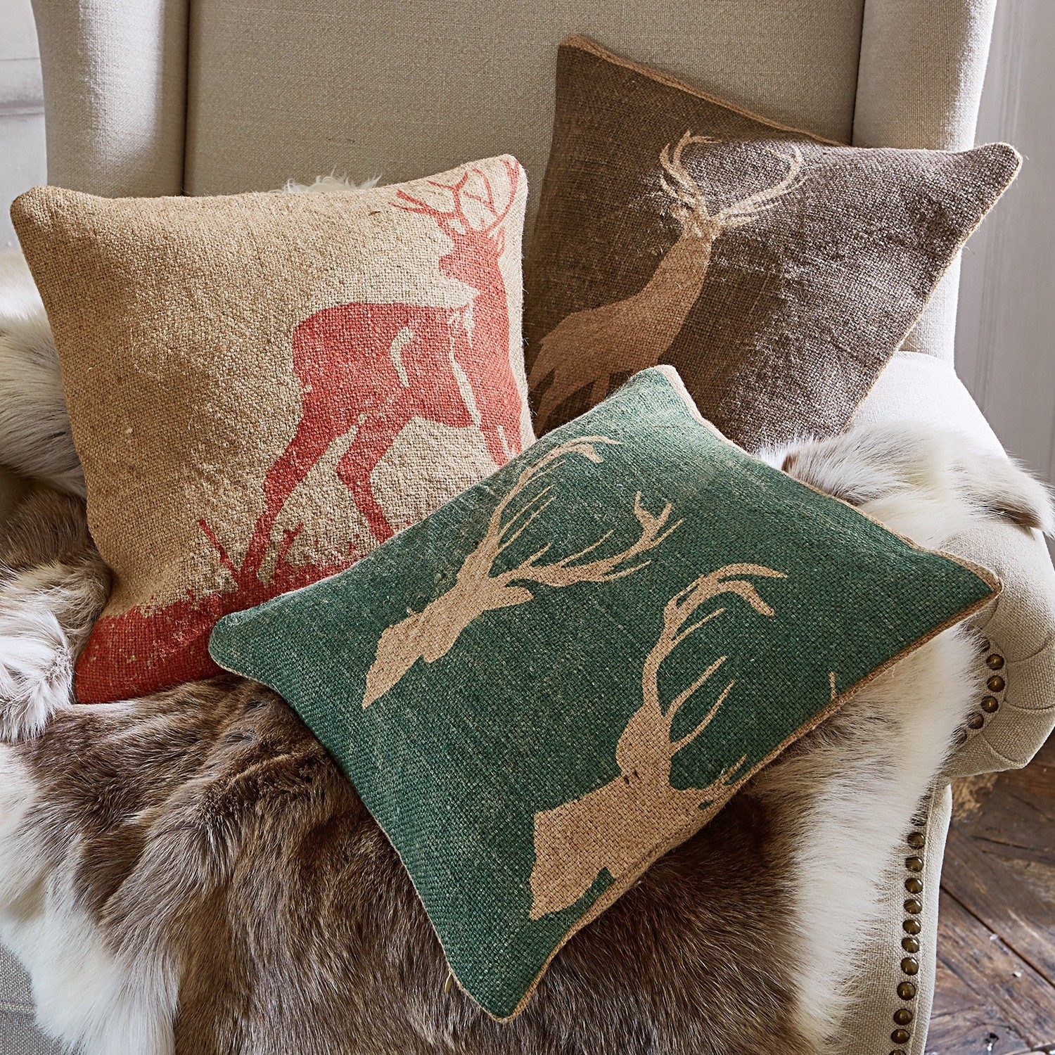 Set de 3 fundas de cojín Shrewton Tres cojines decorativos con motivo de ciervo en colores naturales sobre sillón con manta de piel