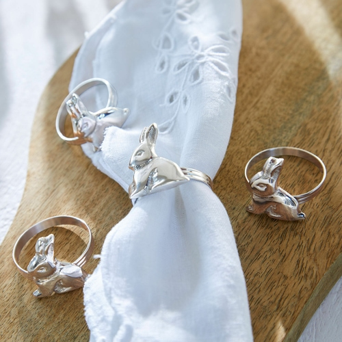 Anillos plateados con diseño de conejo sobre servilleta blanca en tabla de madera, decoración de mesa