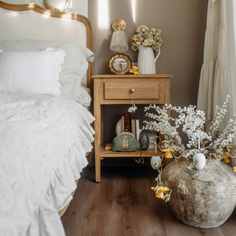 Mesita de noche de madera clara con decoraciones y ropa de cama blanca en dormitorio acogedor de estilo natural
