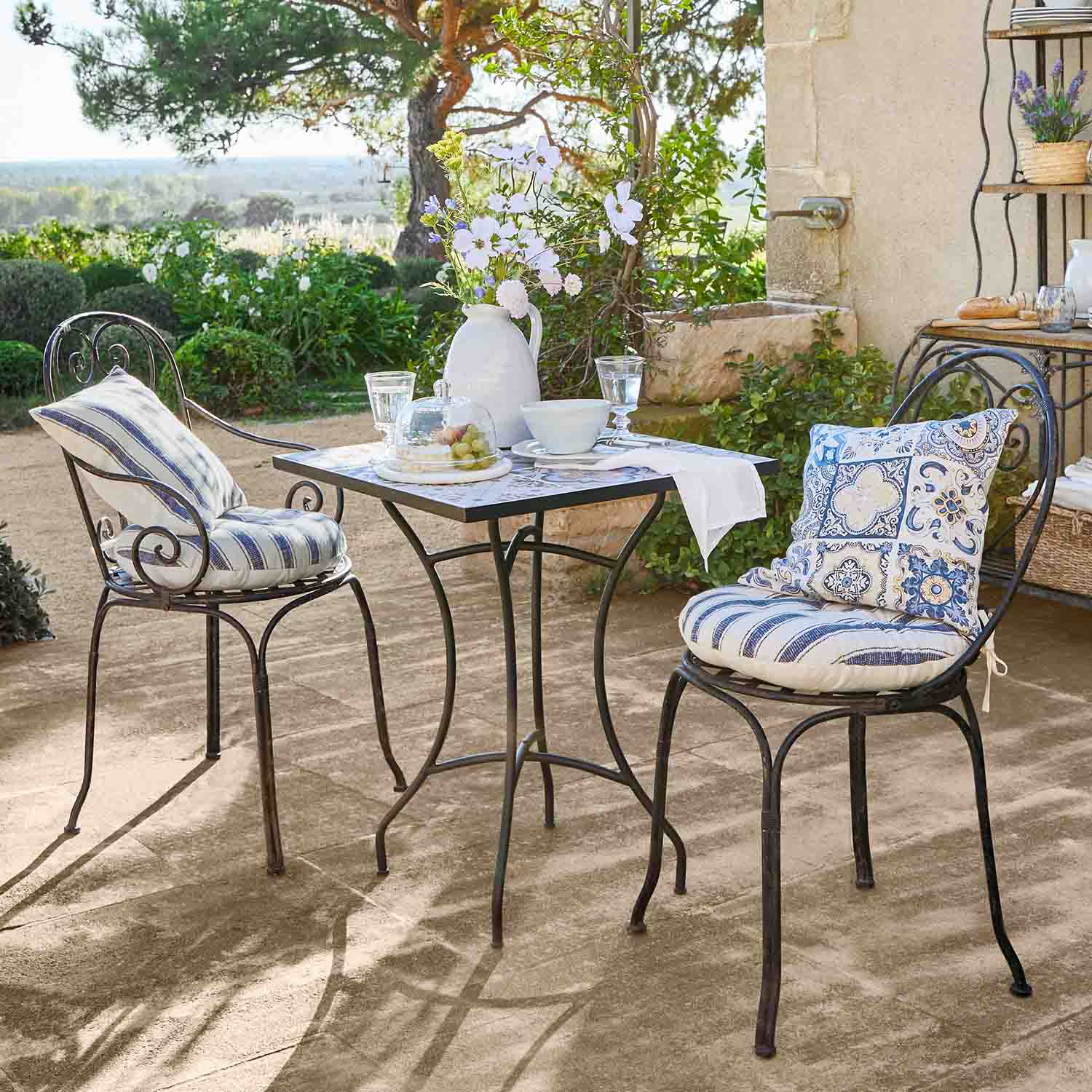 Mesa metálica cuadrada con dos sillas con cojines azules en terraza soleada de jardín