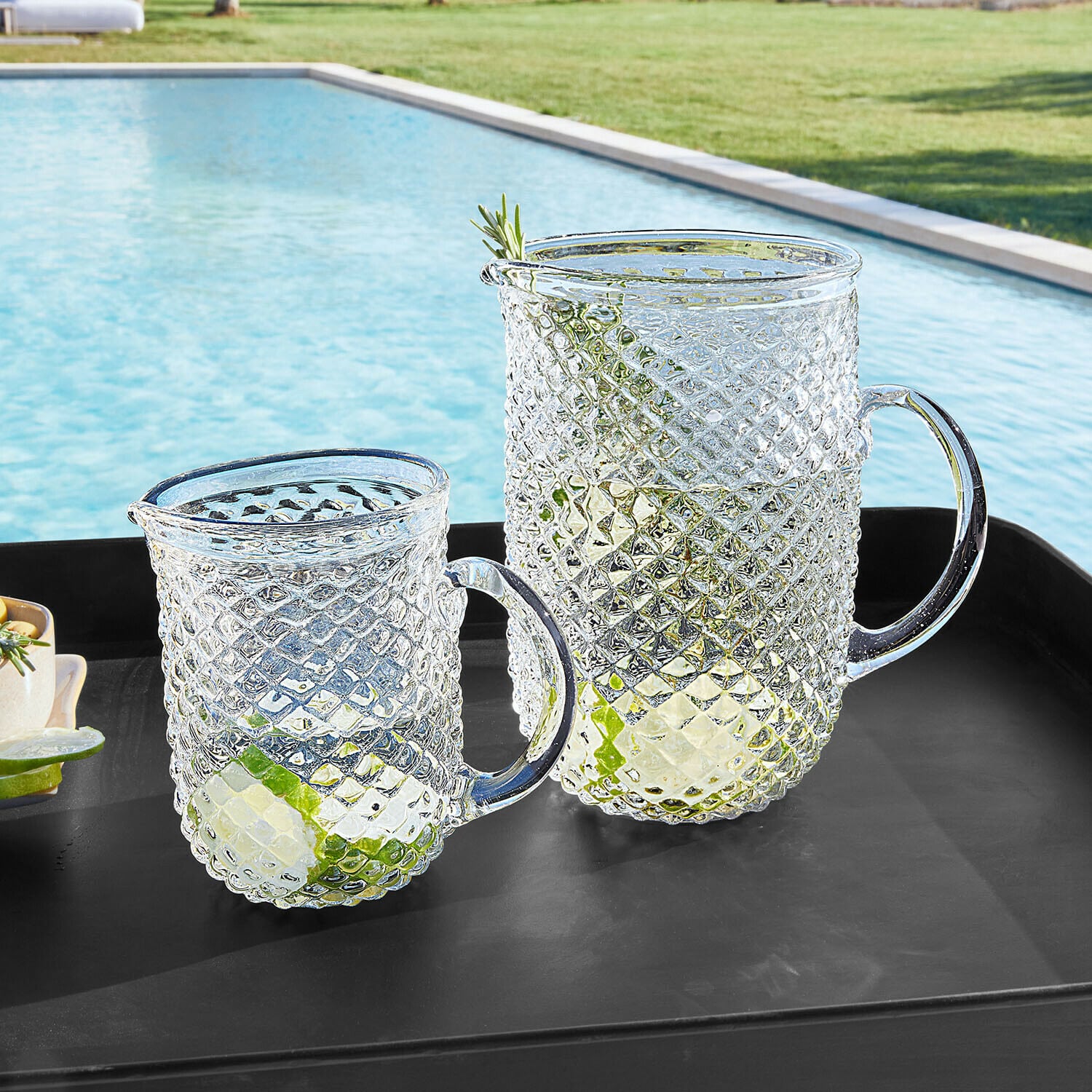 Set de 2 jarras Ivorias Jarra y vaso de vidrio transparente con patrón de diamante, con rodajas de lima, sobre bandeja negra junto a la piscina