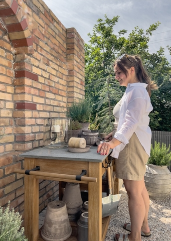 Mujer con camisa blanca y pantalones cortos beige junto a mesa de jardín de madera con encimera de hormigón frente a pared de ladrillo y plantas