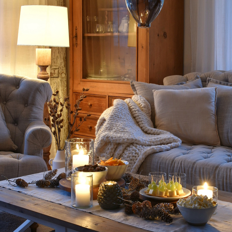 Sala acogedora con manta de punto beige y mesa de madera decorada con velas y aperitivos
