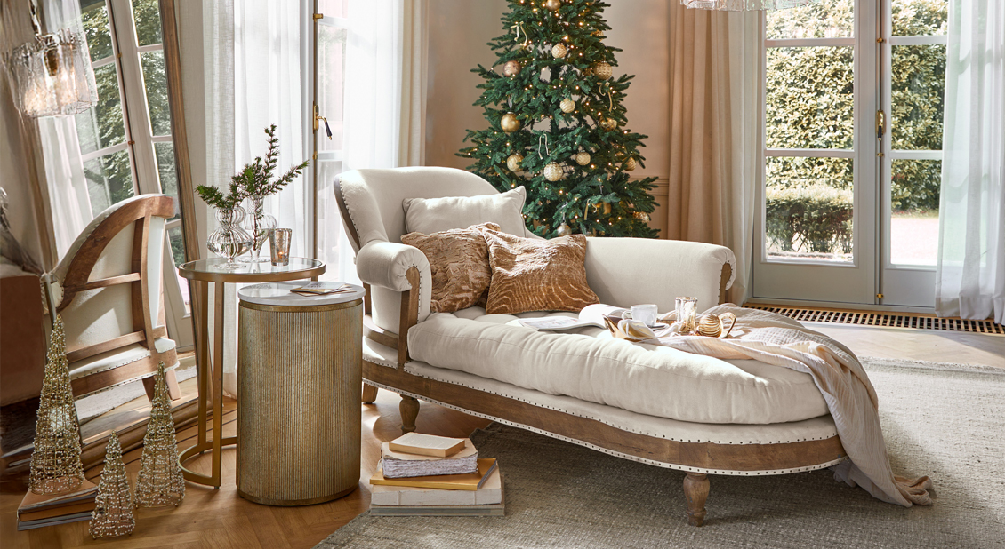 Chaiselongue crema elegante con marco de madera y cojines junto a decoraciones doradas y árbol de Navidad en sala luminosa