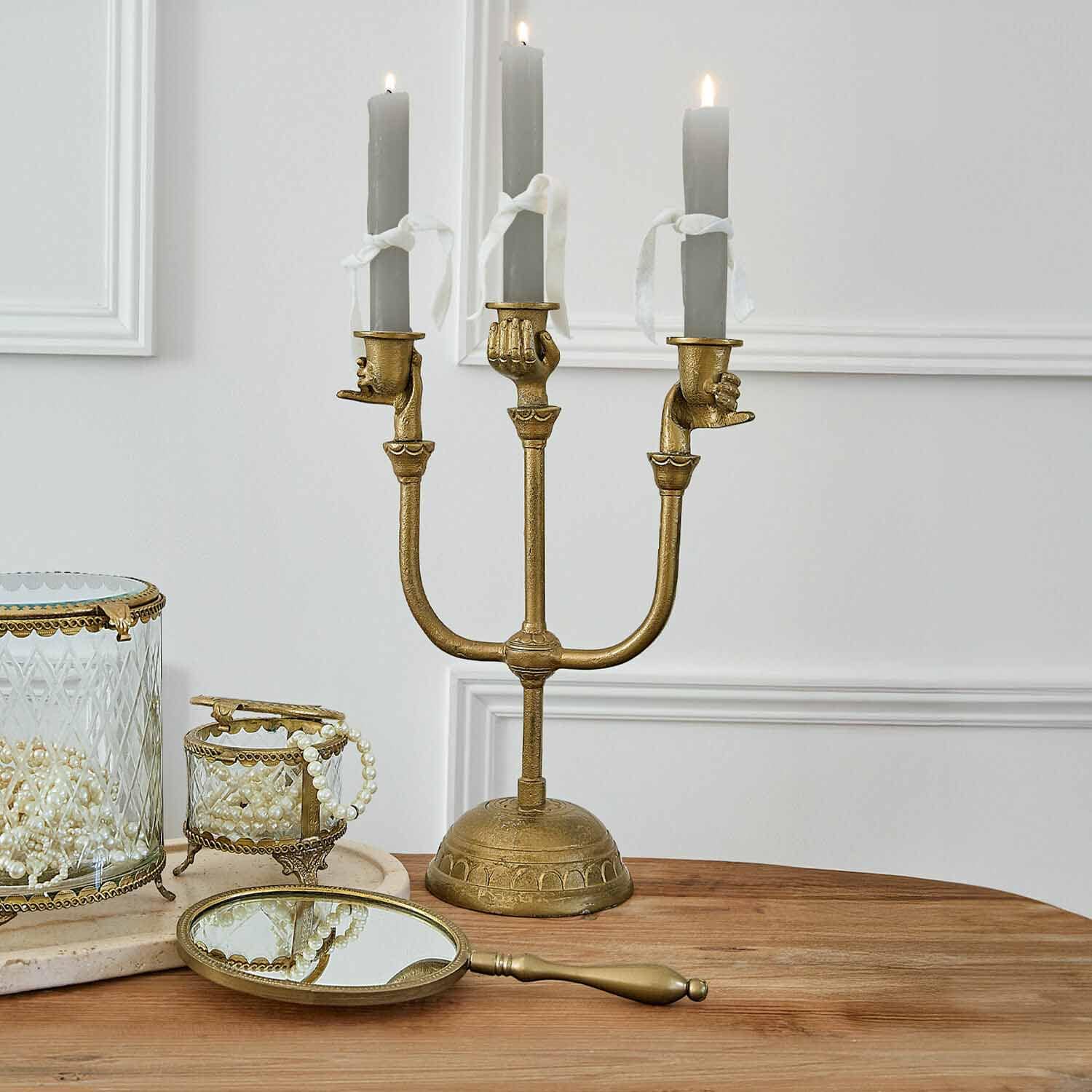 Candelabro dorado con tres velas grises sujetas por manos, decoración de mesa con espejo y cajas de joyas