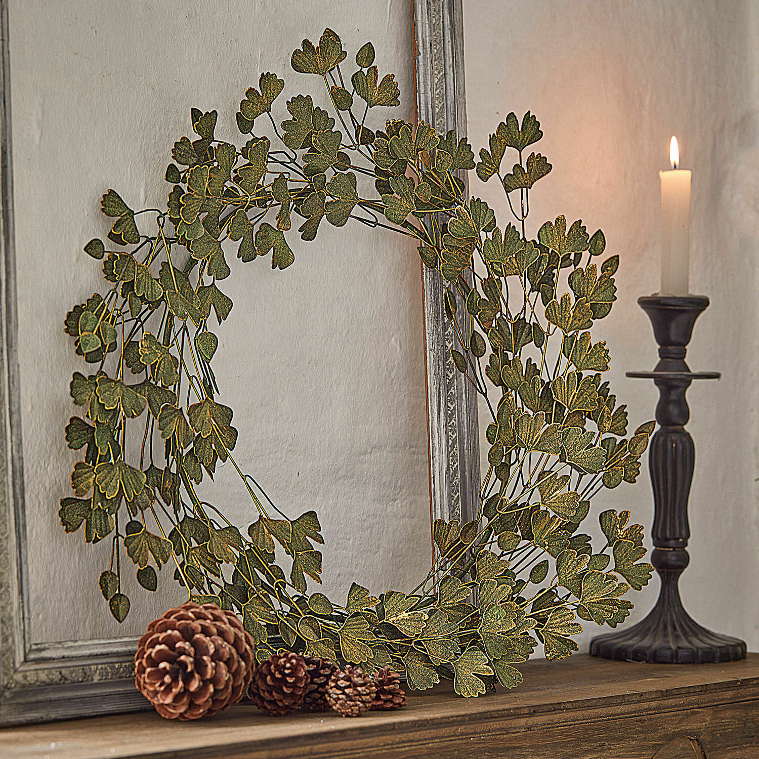 Corona Syrdarja Corona decorativa de hojas verdes sobre estante de madera con piñas y vela encendida