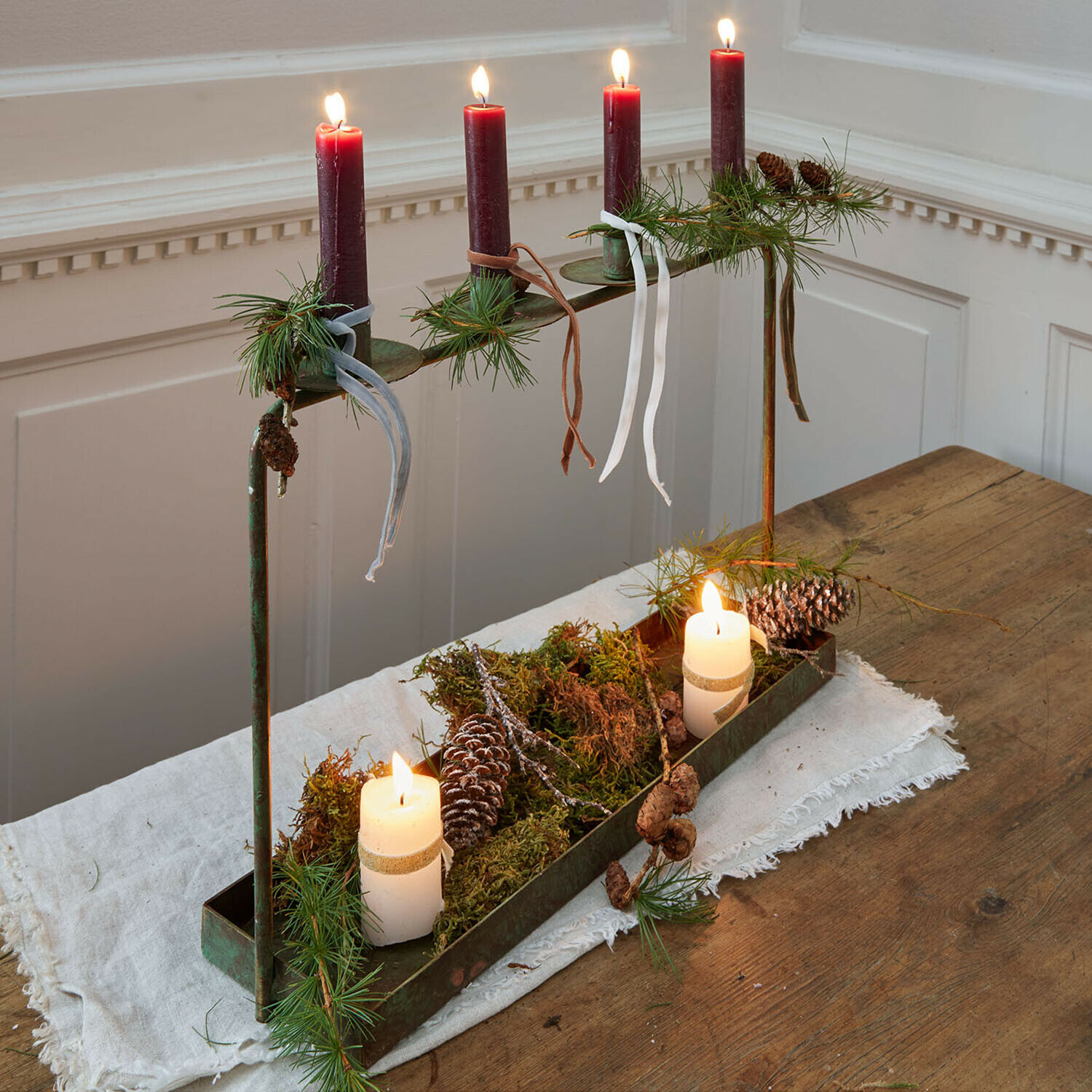 Portavelas Valenzas Candelabro de metal con cuatro velas rojas y decoración de piñas y ramas verdes sobre mesa de madera