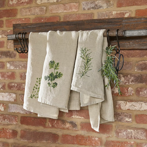 Paños de cocina de lino color natural con estampado de hierbas colgados en soporte de madera frente a pared de ladrillo