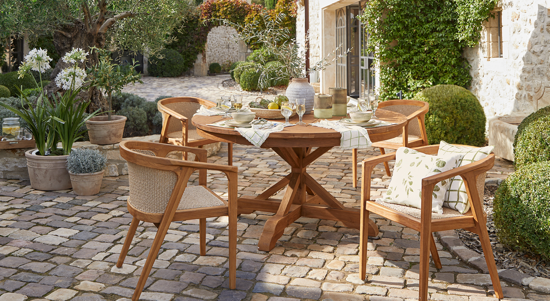 Mesa redonda de madera con cuatro sillas en patio soleado con pavimento de piedra y plantas