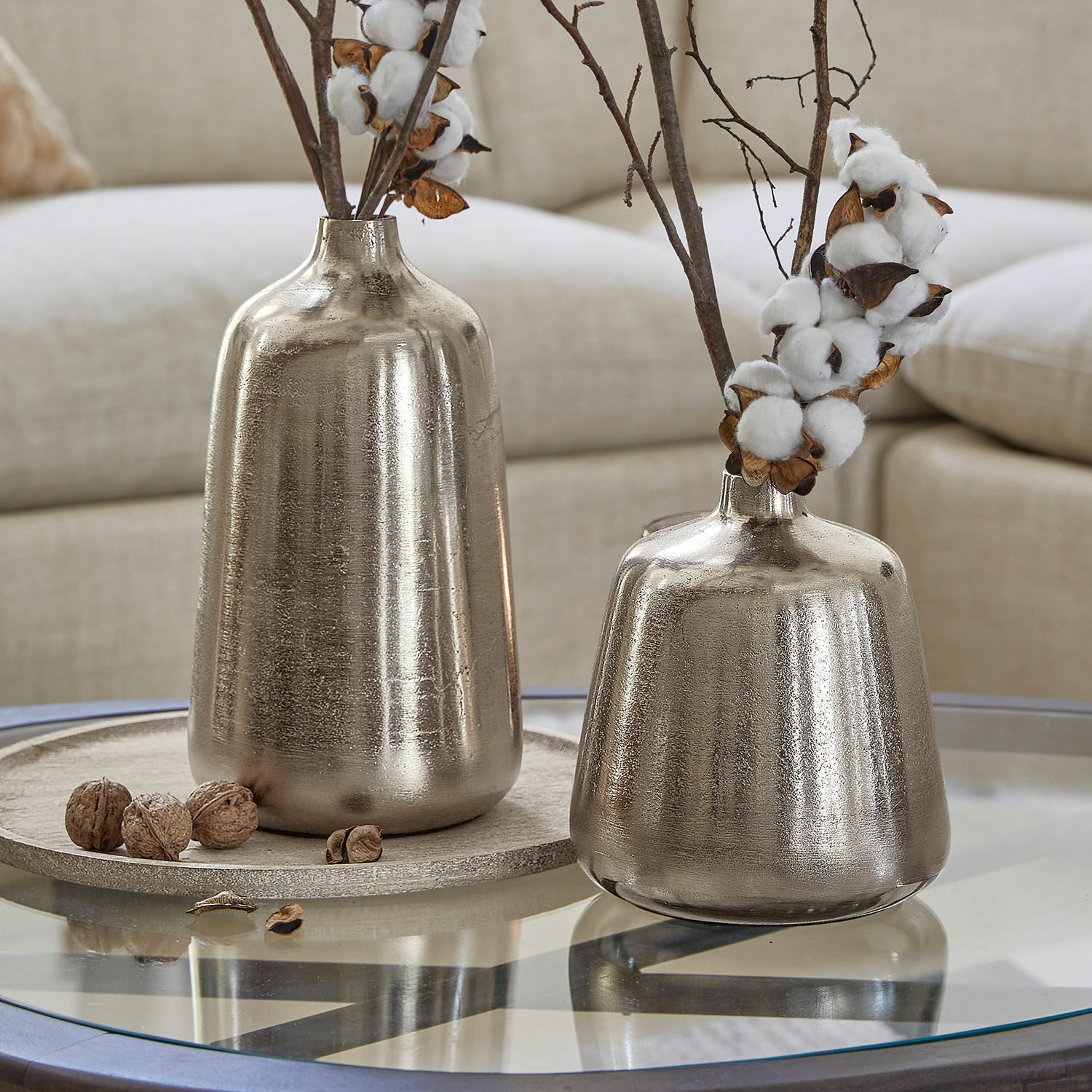 Set de 2 jarrones Orabela Dos jarrones metálicos plateados sobre mesa de cristal con ramas de algodón decorativas y nueces