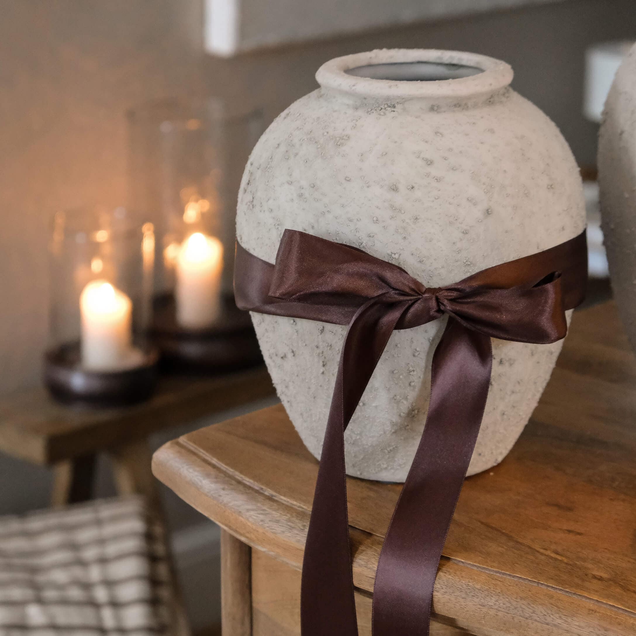 Jarrón decorativo blanco con textura y lazo de satén marrón sobre mesa de madera en ambiente acogedor
