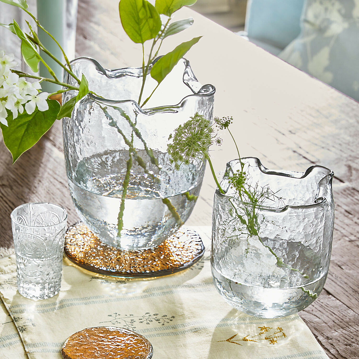 Set de 2 jarrones Felucia Jarrones de vidrio transparente con borde ondulado sobre mesa de madera con plantas verdes y decoración