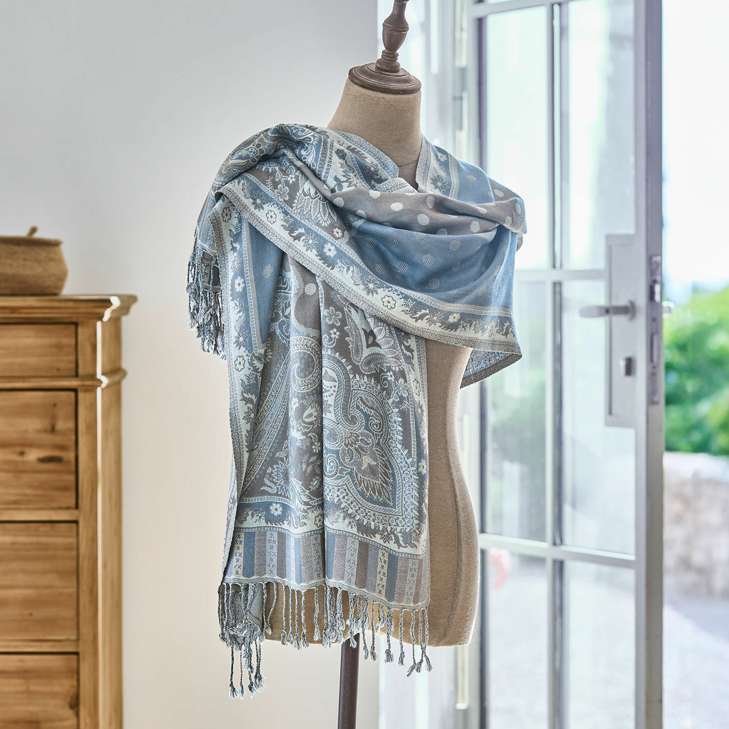 Chal azul grisáceo con diseño paisley y flecos sobre maniquí en habitación luminosa