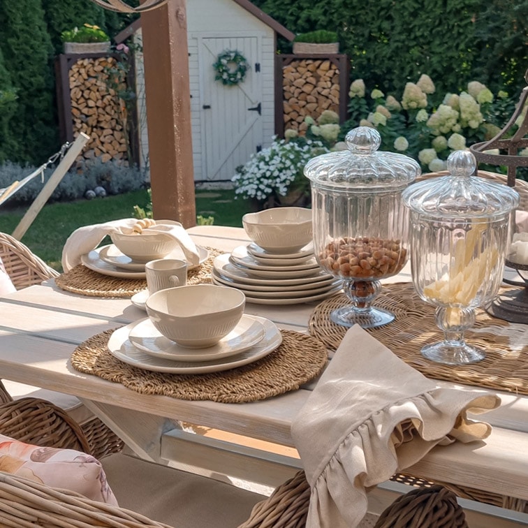 Juego de cerámica blanca sobre mesa de madera clara con frascos de vidrio decorativos en jardín