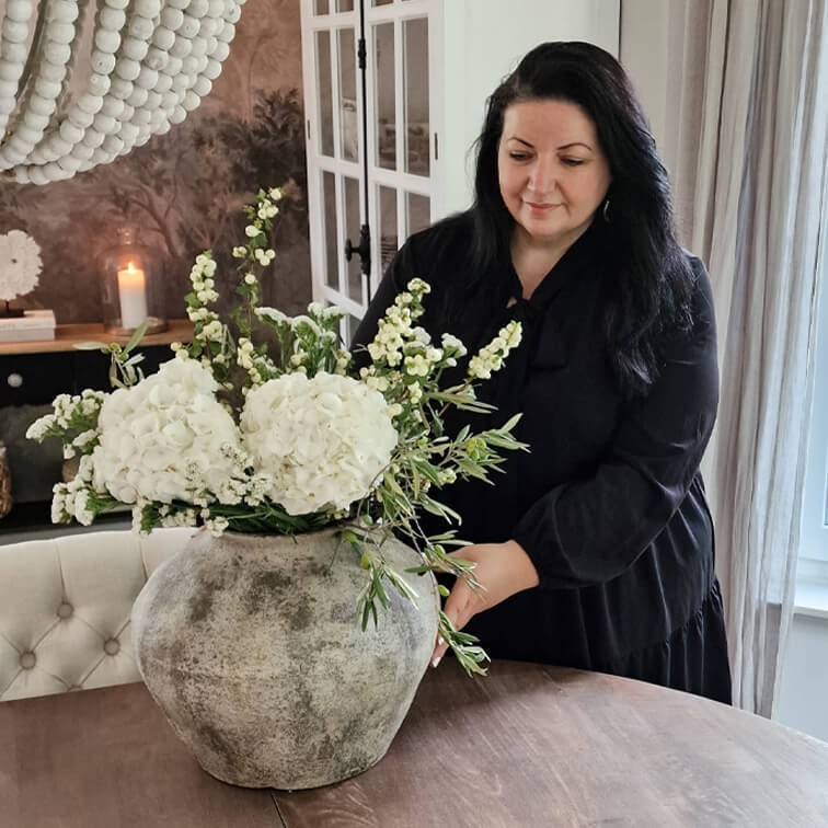 Jarrón grande de cerámica gris con flores blancas sobre mesa de madera en interior acogedor.
