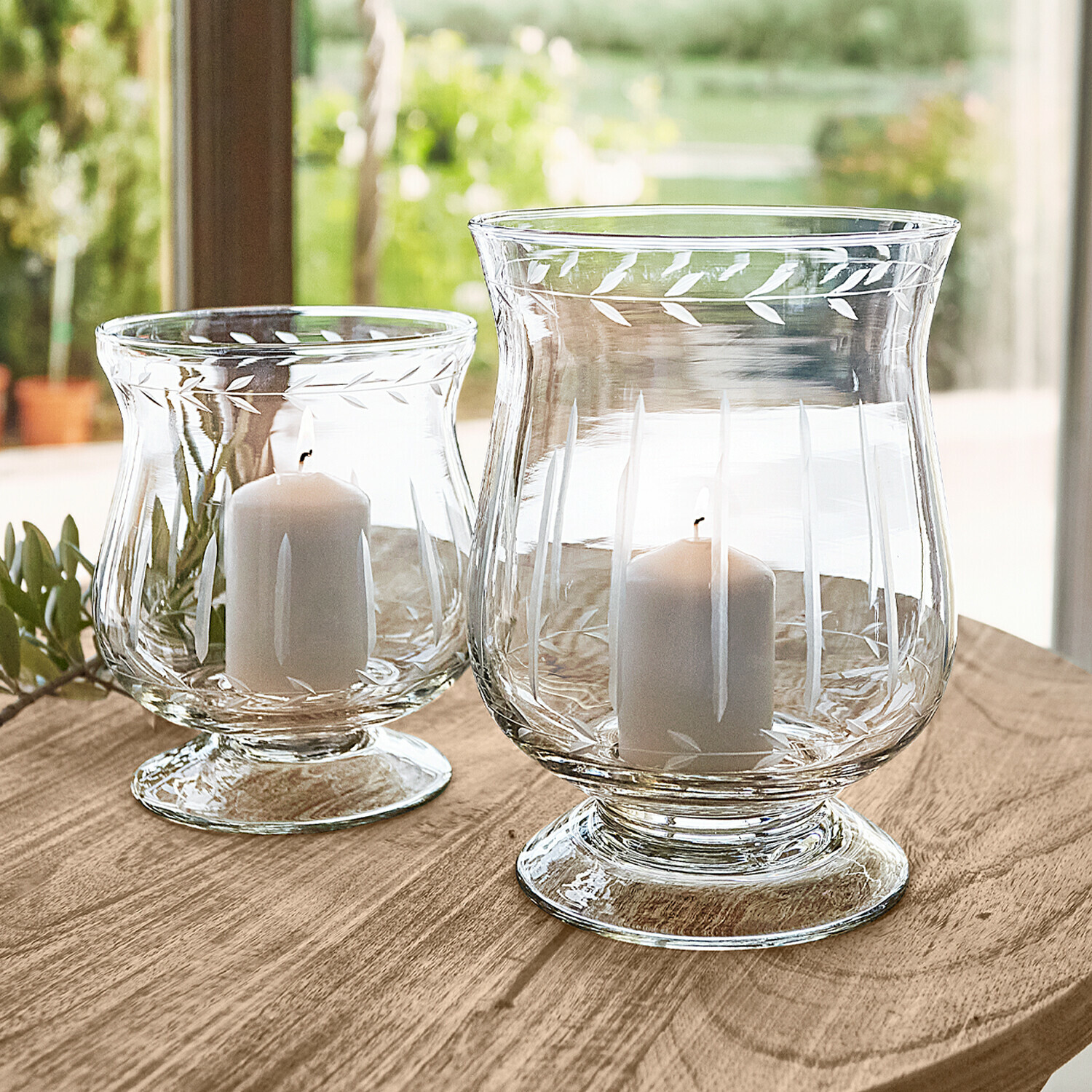 Dos portavelas de vidrio transparente con velas blancas sobre mesa de madera, decoración hojas