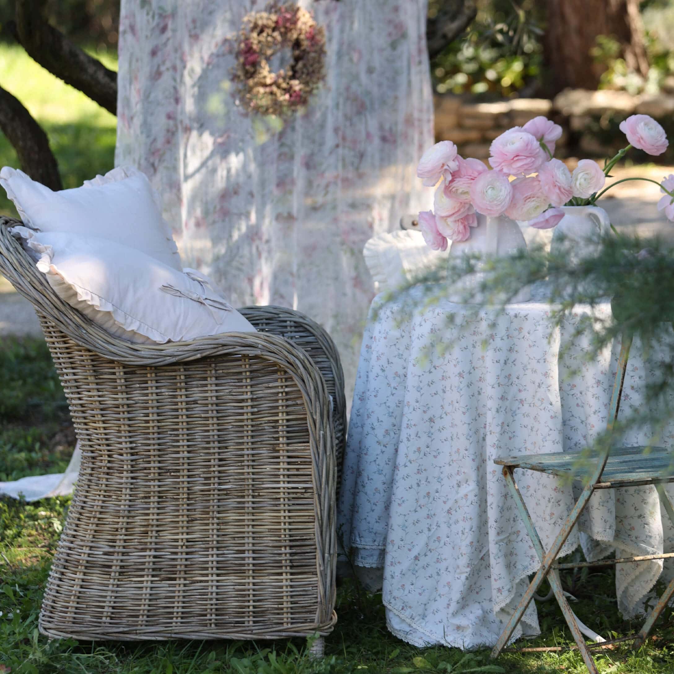 Sillón de mimbre con cojines blancos junto a mesa con mantel floral y flores rosas en jardín
