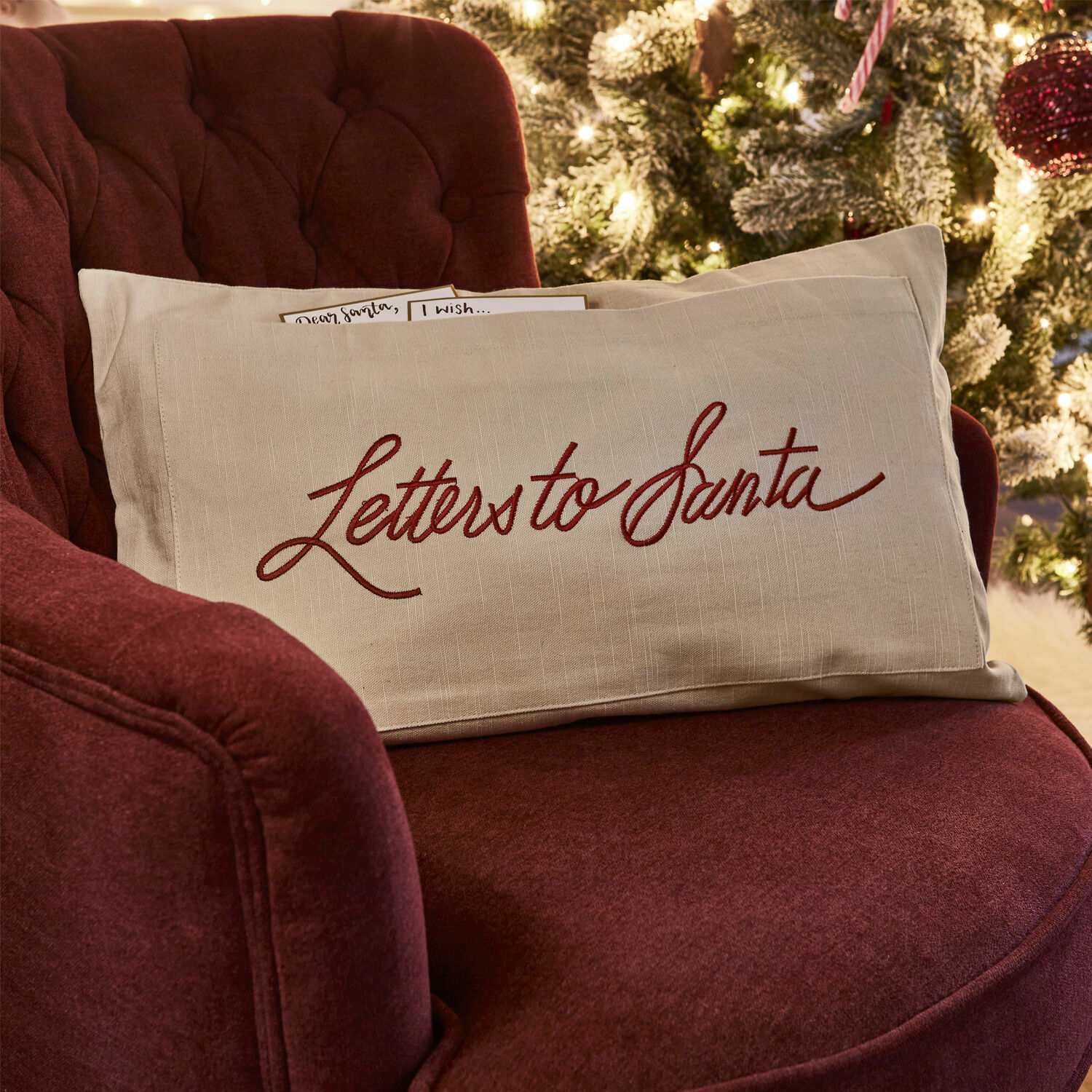 Funda de cojín Welenzas Cojín beige de lino con texto rojo Letters to Santa sobre sillón burdeos junto a árbol de Navidad