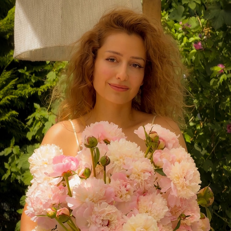 Mujer con cabello largo sosteniendo un gran ramo de peonías rosas frente a un fondo de jardín verde