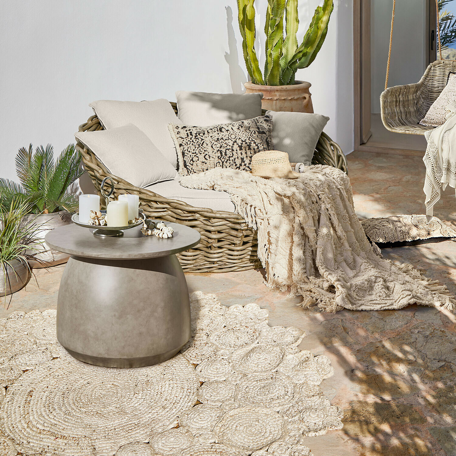 Mesa de centro redonda de hormigón en terraza con sofá de mimbre y manta decorativa crema