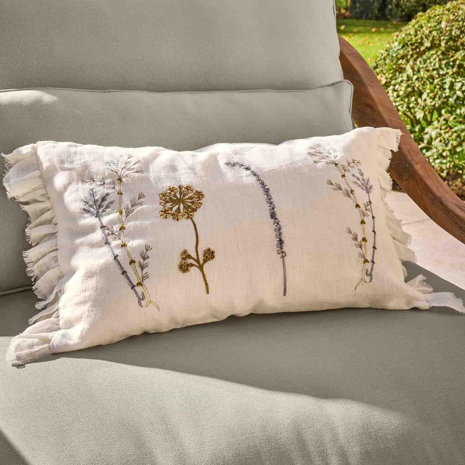 Cojín rectangular de lino blanco con bordado floral y volante sobre sillón gris de jardín