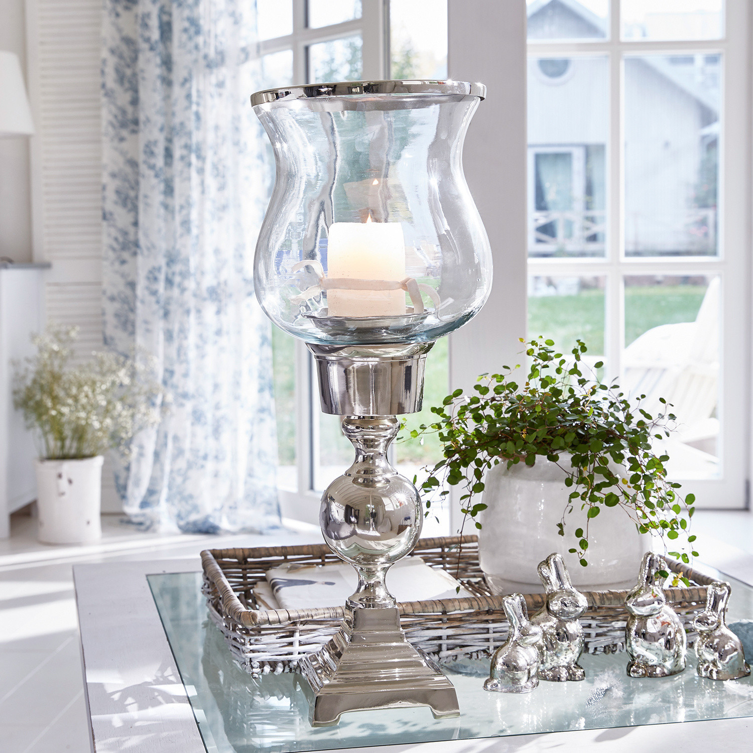 Portavelas de vidrio con base metálica plateada sobre mesa con decoraciones de conejos plateados