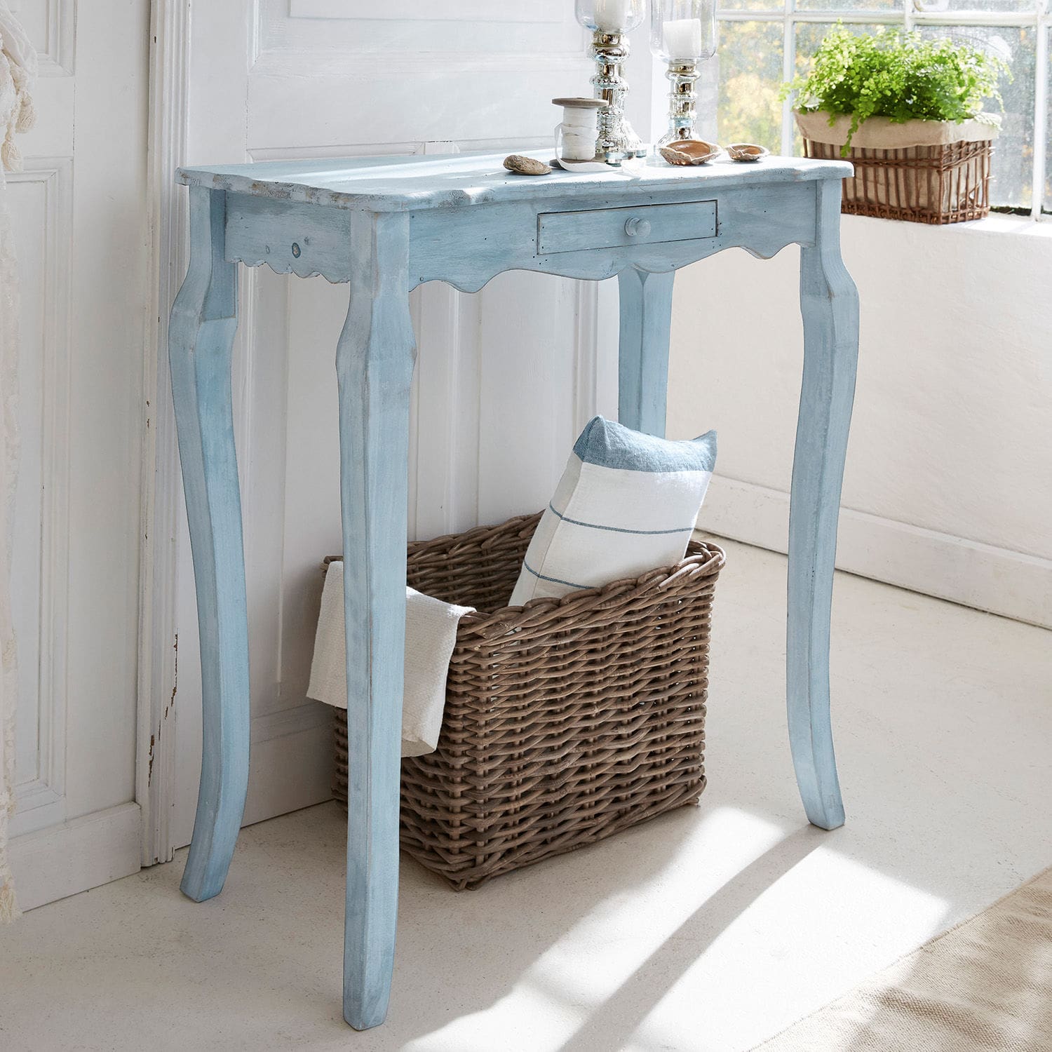 Consola Barbey Mesa azul de madera estilo vintage con cajón y cesta de mimbre, decorada junto a ventana luminosa