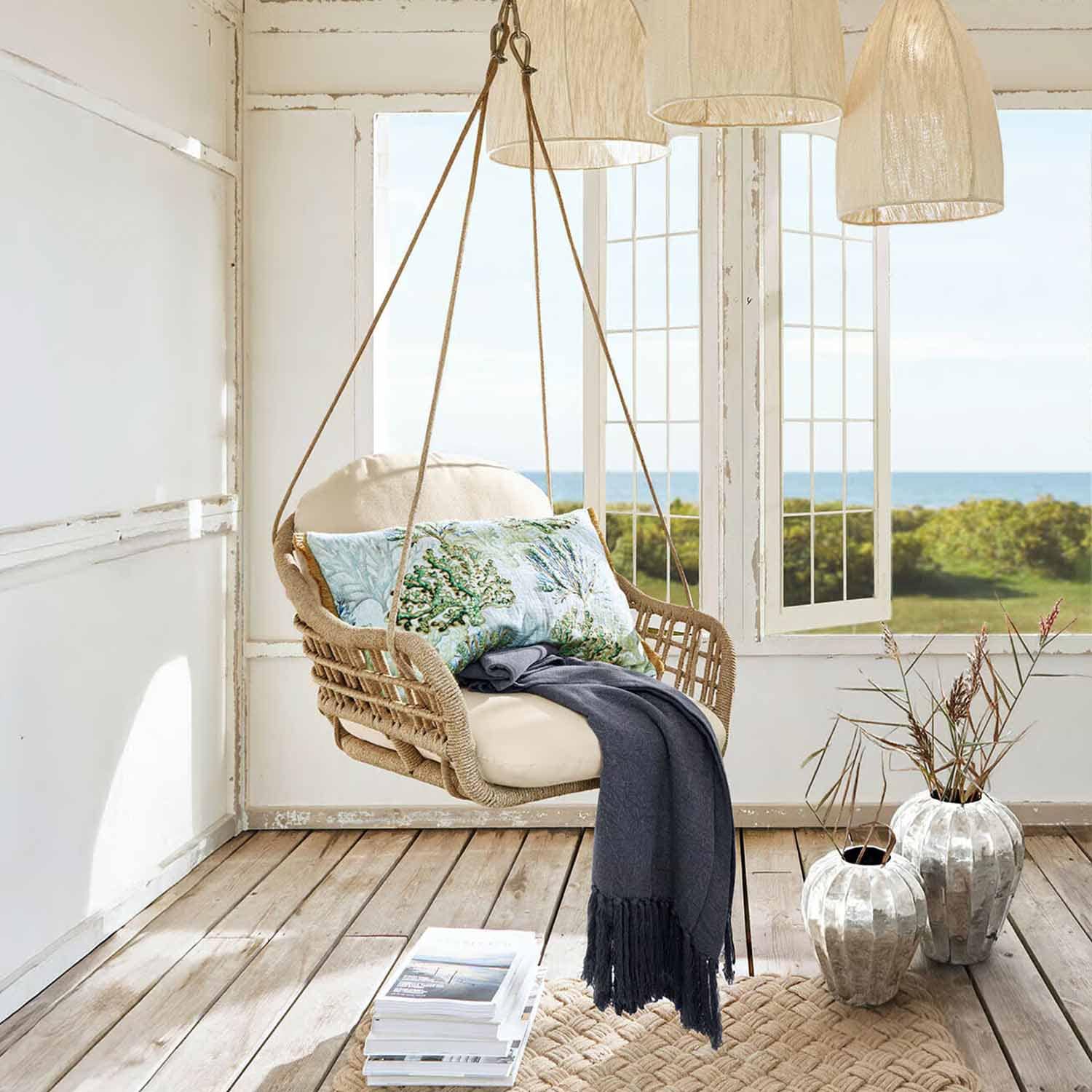 Sillón colgante de cuerda trenzada color natural con cojín crema y manta azul oscuro en habitación luminosa